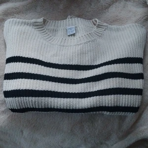 Randig stickad tröja från Lindex - Säljer en ribbstickad tröja från Lindex i beige med svarta horisontella ränder. Tröjan har rund halsringning och långa ärmar. Perfekt för dig som gillar en klassisk och chill stil. Materialet är mjukt och skönt, passar bra till jeans eller kjol.