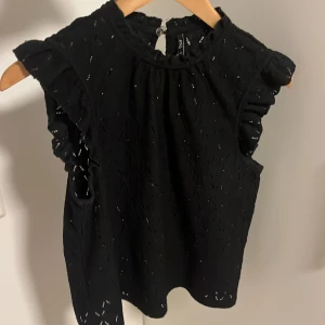 Svart blus med volang från ONLY - Supersöt svart blus från ONLY i storlek XS. Blusen har broderade hål, volangdetaljer vid ärmarna och en liten knapp i nacken. Perfekt för dig som gillar romantiska och feminina detaljer. Tunn och luftig känsla.