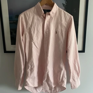 Rosa- och vitrandig skjorta Ralph Lauren - Säljer en klassisk rosa- och vitrandig skjorta från Ralph Lauren i bomull. Skjortan har lång ärm, knappar framtill och broderad logga på bröstet. Custom fit-modell med krage och snygg passform. Perfekt för dig som gillar stilrena och tidlösa plagg.
