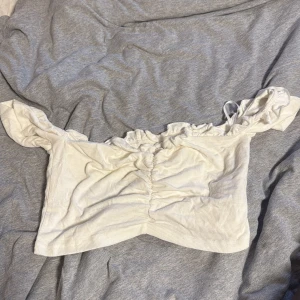 Vit offshoulder croppad topp - Supersöt vit croppad topp med offshoulder och rynkade detaljer framtill. Toppen har volangkant upptill och är tillverkad i ett mjukt, ribbat material. Perfekt för en somrig look med lite romantisk vibe.