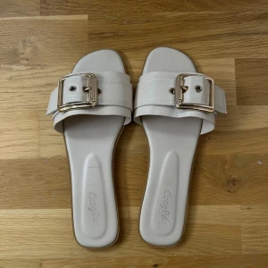 Vita sandaler med stor spänne - Snygga vita sandaler med bred rem och stor guldfärgad spänne. Platt sula och fyrkantig tå ger en modern vibe. Perfekta för varma dagar när du vill ha en clean och stilren look. Lite stora i storleken