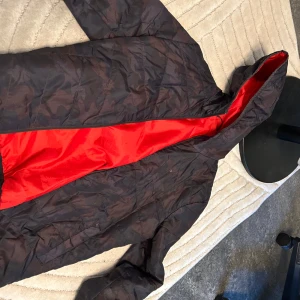 Svart och röd vindjacka med luva - Snygg svart vindjacka med röd insida och luva. Jackan har en lätt och smidig känsla, perfekt för blåsiga dagar. Stängs med dragkedja och har en sportig look. Passar dig som gillar att sticka ut med färgdetaljer.