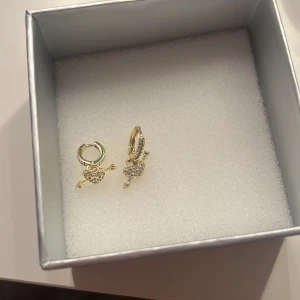 Guldiga örhängen med hjärthänge - Säljer ett par snygga guldiga örhängen med hängande hjärta och pil, dekorerade med små glittriga stenar. Perfekt för dig som vill ha något gulligt och trendigt! Hängena har detaljerad design och ger en extra touch till din look.