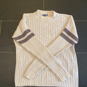 Ribbstickad beige tröja Blount & Pool - Ribbstickad beige tröja från Blount & Pool med lång ärm och två mörkgrå ränder på varje ärm. Tröjan har rund halsringning och är i ett mjukt, stickat material som känns både klassiskt och chill.