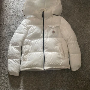 Vit dunjacka från Moncler med huva - Säljer en riktigt snygg vit dunjacka från Moncler med avtagbar huva och klassisk logga på bröstet. Jackan har dragkedja framtill, fickor med dragkedja och en extra ficka med knapp på bröstet. Perfekt för kalla dagar när du vill hålla stilen.