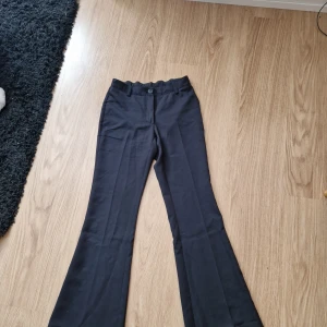 Svarta bootcut kostymbyxor RYVLS - Svarta kostymbyxor från RYVLS i storlek 164. Byxorna har bootcut-modell med utsvängda ben, bälteshällor och två fuskfickor bak. Snygg och stilren look, perfekta till en trendig outfit.