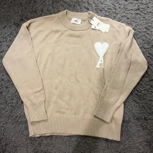 Beige stickad tröja från Ami - Snygg beige stickad tröja från Ami med rund hals och ribbade muddar. På bröstet finns ett vitt hjärta med bokstaven A, vilket ger en unik touch. Perfekt för dig som gillar stilrena och trendiga plagg.