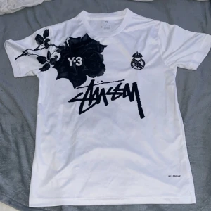 Vit Y-3 x Stüssy Real Madrid t-shirt - Snygg vit t-shirt med svart rosprint fram och bak, Y-3 och Real Madrid-logga samt Stüssy-grafik på bröstet. Tillverkad i lätt polyester med Aeroready-teknologi. Perfekt för dig som gillar streetwear och unika samarbeten.