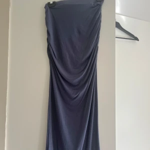 Mörkblå/mörkgrå långkjolmed drapering - Säljer en mörkgrå långkjol med snygg drapering längs sidorna. Kjolrnhar en tight passform och är axelbandslös, vilket ger en cool och elegant siluett. Materialet känns mjukt och stretchigt, perfekt för dig som gillar att sticka ut.