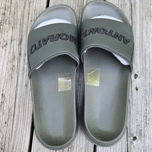 Antony Morato grå/gröna badsandaler - Coola slides från Antony Morato i en mörkgrön färg med svart logotyp över foten. Tillverkade i syntetmaterial med bred rem och räfflad sula för extra grepp. Perfekta för sommardagar vid poolen eller stranden. Storlek 41, tofflor är nya, men utan kartong. Mycket bekväma.