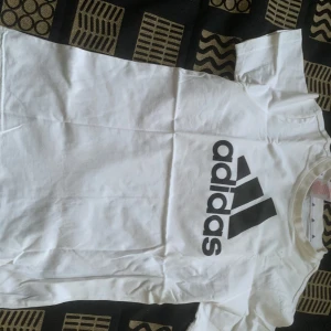 Vit Adidas t-shirt - Fräsch vit t-shirt från Adidas med stor svart logga framtill. Klassisk rund hals och korta ärmar. Tillverkad i mjuk bomull som är skön mot huden. Perfekt för dig som gillar sportig och enkel stil.