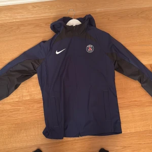 PSG Nike blå vindjacka med huva - Snygg blå vindjacka från Nike med PSG-logga på bröstet. Jackan har svarta detaljer på axlar och ärmar, dragkedja framtill och praktisk huva. Perfekt för dig som gillar fotboll och vill ha en sportig stil. Materialet är lätt och känns skönt att bära på sig.