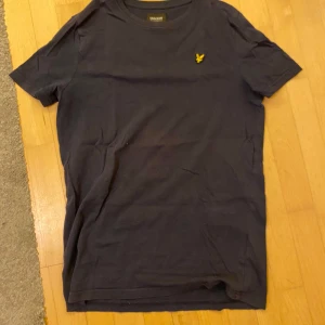 Mörkblå t-shirt från Lyle & Scott - Snygg mörkblå t-shirt från Lyle & Scott med klassisk gul logga på bröstet i storlek S. T-shirten är i mjuk bomull och har en normal passform med korta ärmar. Perfekt till jeans eller shorts för en clean och enkel stil. Pris kan diskuteras!