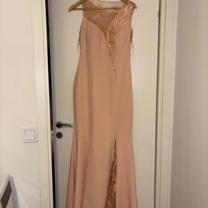 Champagnefärgad långklänning med broderi - Säljer en elegant långklänning i champagnefärg med transparent mesh upptill och detaljerat broderi i samma ton. Klänningen har en v-ringad front, slits framtill och är ärmlös. Perfekt för bal, bröllop eller fest, med en lyxig och sofistikerad vibe.