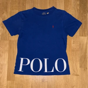 Blå Polo Ralph Lauren t-shirt - Blå t-shirt från Polo Ralph Lauren med stor vit POLO-text nedtill och klassisk röd broderad logga på bröstet. T-shirten är kortärmad och gjord i mjuk bomull, perfekt för en clean och stilren look. Priset kan diskuteras! Skriv vid minsta fråga!