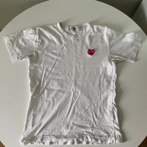 Vit T-Shirt från CDG Play - Stl S - Vit T-shirt av märket comme des garçons (cdg) play i bra skick. De enda små skavankerna är att den har en pytteliten orange fläck på framsidan men om ni kollar på första bilden så märker ni att det inte är nåt som syns, och att nedre sömmen har börjat släppa lite, ingenting som syns för någon annan! Köpt på NK för 799. Tveka inte att höra av er med prisförslag eller funderingar!