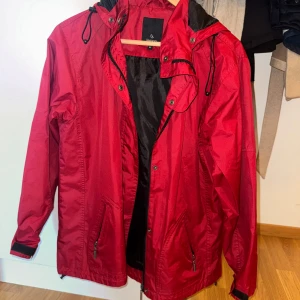 Röd vind/regnjacka från Lemon, storlek M - Röd vind/regnjacka från Lemon med huva och svarta detaljer. Jackan har dragkedja framtill, två fickor med dragkedja och justerbar snörning i huvan. Perfekt för blåsiga dagar och enkel att matcha med din stil.