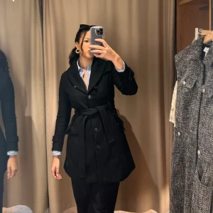 Stilren ull  svart kappa med bälte, bakslit och knappar! - Stilren svart kappa från Saint Tropez med markerad midja tack vare bälte, stora knappar och bred krage. Långärmad modell i ullblandning, perfekt för kyliga dagar. Snygg och tidlös design som funkar till många outfits.