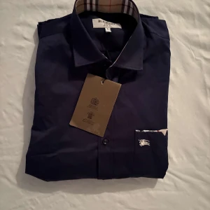 Mörkblå skjorta från Burberry - Snygg mörkblå skjorta från Burberry med klassisk krage och knappar framtill. Ikoniskt rutigt mönster på insidan av kragen och liten broderad logga på bröstfickan. Perfekt för dig som gillar stilrena och exklusiva plagg. .     Nypris: 7449kr GORILLAPRIZE:2915kr             Skick:10/10 oöppnad          KVITTO MEDKOMMER 