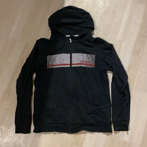 Svart hoodie från Boss med dragkedja - Svart hoodie från Boss med hel dragkedja och huva. Framsidan har ett rutigt mönster i vitt och rött med Boss-loggan. Tröjan är långärmad och har en klassisk passform, perfekt för en chill och stilren look.