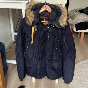 Parajumpers mörkblå dunjacka S - Mörkblå Parajumpers dunjacka med päls på huvan, flera fickor och snygga detaljer som gul rem och patch på ärmen. Jackan har dragkedja, knappar och är perfekt för kalla dagar. Klassisk vinterjacka med cool look och funktionella detaljer. Pris kan diskuteras 