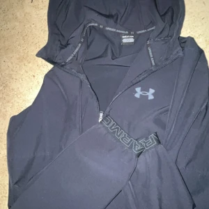 Svart vindjacka från Under Armour - Svart vindjacka från Under Armour med stilren design och logga på bröstet. Jackan har hel dragkedja framtill, två sidofickor med dragkedja och långärmade ärmar. Perfekt för aktiva dagar och sportiga outfits.