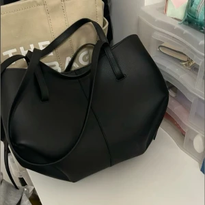 polene liknande väska - Snygg svart axelväska med stilren design och mjukt material. Väskan har två långa handtag så du kan bära den både på axeln och i handen. Perfekt storlek för att få plats med allt du behöver till skolan eller stan.