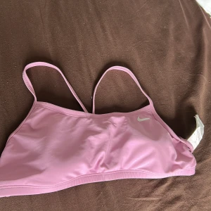 Ljusrosa linnetopp från Nike - Säljer en ljusrosa linnetopp från Nike med tunna axelband och enkel design. Toppen är tillverkad i ett mjukt och stretchigt syntetmaterial som passar perfekt för träning eller sportiga outfits. Nike-loggan syns framtill i vitt.