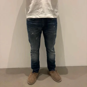 Blå jeans med slitningar - Säljer ett par blå jeans med snygga slitningar och tvättade detaljer. Jeansen har klassisk femficksdesign och raka ben. Perfekta för en avslappnad och trendig look. Passar dig som gillar en lite tuffare stil.