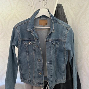 Ljusblå jeansjacka från 157 - Snygg ljusblå jeansjacka från 157 med klassisk krage, knappar framtill och två bröstfickor med lock. Jackan har en rak passform och är tillverkad i slitstarkt denim. Perfekt att slänga över en hoodie eller t-shirt.