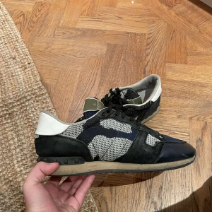 Valentino Rockrunner sneakers blå/svart - Säljer ett par Valentino Rockrunner sneakers i blått, svart och vitt med coolt kamouflagemönster. Skorna har snörning, platt sula och detaljer i mocka och textil. Perfekta för dig som gillar exklusiva och trendiga sneakers med unik design.