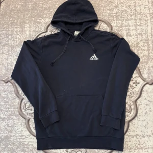 Mörkblå  hoodie från Adidas med huva - Klassisk Mörkblå hoodie från Adidas med vit logga på bröstet. Tröjan har huva med snörning, känguruficka och långa ärmar. Perfekt för en chill och sportig stil. Tillverkad i mjuk bomullsmix som känns skön mot huden.