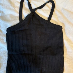 Svart ribbad linnetopp med korsade band - Säljer en svart ribbad linnetopp med smala axelband som korsas i framtill. Toppen är croppad och har en tight passform, perfekt till jeans eller kjol. Materialet är mjukt och stretchigt, vilket gör den superbekväm att bära.