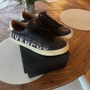 Givenchy svarta sneakers med logga - Svarta sneakers från Givenchy med stor vit logga längs sidan och vit hälkappa med Givenchy Paris-tryck. Skorna har svarta skosnören, vit sula och är tillverkade i skinn. Klassisk rund tå och låg modell som ger en clean och lyxig vibe. Hör av er vid frågor 