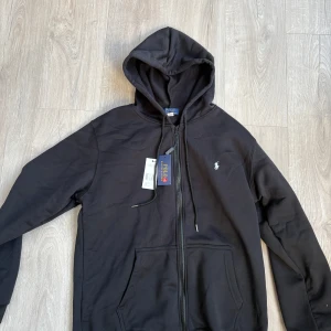 Svart hoodie från Polo Ralph Lauren - Svart hoodie från Polo Ralph Lauren med dragkedja och huva. Klassisk design med vit broderad logga på bröstet och känguruficka framtill. Mjuk insida och snörning i huvan. Perfekt för en chill och stilren look.
