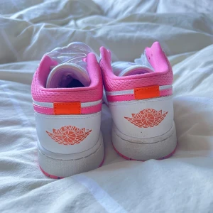 Nike Air Jordan 1 Low vit/rosa - Säljer ett par Nike Air Jordan (tror 1 Low) sneakers i vitt och rosa med orange detaljer. Skorna har klassisk snörning, perforerad tå och rosa swoosh på sidan. Använt fåtal gånger. De saknar dock ett skosnöre på ena skon men går lätt att ersätta med ett vitt snöre.