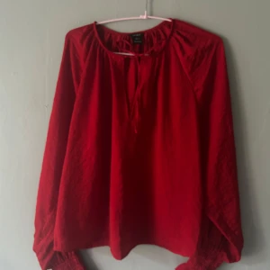 Röd blus med knyt från Lindex - Säljer en röd blus från Lindex i storlek S. Blusen har långa ärmar med smockade muddar och en snygg knytning vid halsen. Luftig och flowy passform, perfekt till jeans eller kjol. Materialet känns lätt och bekvämt.