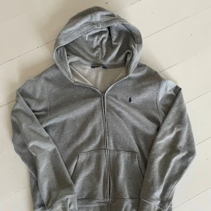 Grå hoodie från Polo Ralph Lauren - Mysig grå hoodie från Polo Ralph Lauren med dragkedja och huva. Tröjan har mjukt fleecefoder på insidan, två fickor framtill och klassisk broderad logga på bröstet. Perfekt för chill dagar eller när du vill ha en clean och enkel look. Pris kan diskuteras