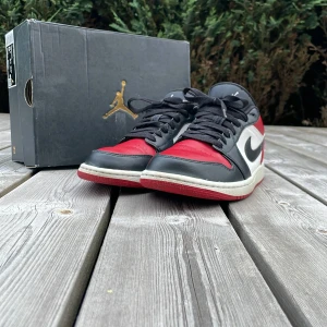 Nike Air Jordan 1 Low röd/svart/vit - Säljer ett par Nike Air Jordan 1 Low med klassisk färgblockning i rött, svart och vitt. Skorna har svart swoosh, snörning och ikoniska Jordan-loggor på plös och häl. Ovandel i skinn och syntet, platt sula och rund tå. Perfekta för dig som vill sticka ut med en tidlös sneaker.