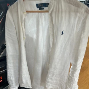 Vit linneskjorta från Polo Ralph Lauren - Snygg vit linneskjorta från Polo Ralph Lauren med klassisk blå logga broderad på bröstet. Skjortan har lång ärm, knappar framtill och en avslappnad passform. Perfekt för en fräsch och stilren look. Jag säljer skjortan då den är för liten för mig just nu.