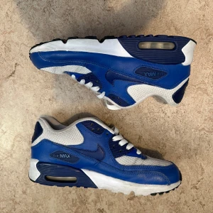 Nike Air Max 90 blå och vita sneakers - Nike Air Max 90 sneakers i blått och vitt med klassisk synlig Air Max-dämpning i sulan. Ovandel i skinn och mesh för både stil och andningsförmåga. Snygga detaljer med vita skosnören och svart yttersula. Perfekta för dig som gillar retrovibbar och streetstyle.