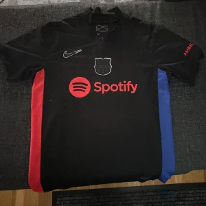 FC Barcelona svart Nike fotbollströja - Säljer en FC Barcelona fotbollströja från Nike med Dri-Fit material. Tröjan är svart med röda och blå sidopaneler, har korta ärmar och V-ringad krage. Spotify-logga på bröstet och UNHCR-tryck på ryggen. Perfekt för dig som älskar Barca och vill ha en snygg matchtröja.