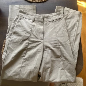 Beiga kostymbyxor från NA-KD, stl 36 - Snygga beiga kostymbyxor från NA-KD i klassisk rak modell med pressveck och hällor för bälte. Byxorna har sidfickor och är tillverkade i ett mjukt bomullsmaterial som känns skönt mot huden. Storlek 36.