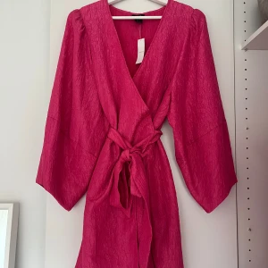 Rosa omlottklänning från Lindex - Säljer en snygg rosa omlottklänning från Lindex i storlek S. Klänningen har en cool textur, breda långa ärmar och ett knytband i midjan. Prislapp sitter kvar. 