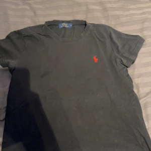 Svart t-shirt från Ralph Lauren - Säljer en klassisk svart t-shirt från Ralph Lauren med det ikoniska röda logotypbroderiet på bröstet. T-shirten är kortärmad och tillverkad i mjuk bomull för en bekväm passform.