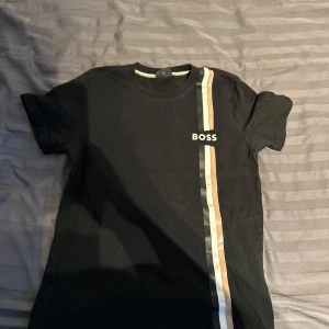 Svart t-shirt från Boss med randig detalj - Snygg svart t-shirt från Boss med en vertikal randig detalj i vitt svart och beige på framsidan. Perfekt för en stilren look. Kortärmad och tillverkad i mjukt material för bekväm passform.