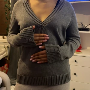 Grå v-ringad tröja med knappar - Säljer en stilren grå tröja med v-ringning och ribbade detaljer. Tröjan har långa ärmar med dekorativa knappar vid ärmsluten. Perfekt för en avslappnad look. 