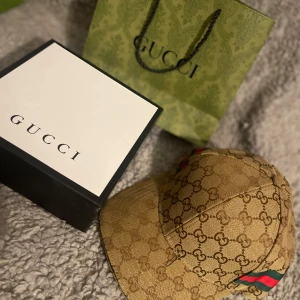 Beige keps från Gucci - Snygg beige keps från Gucci med det ikoniska GG-mönstret. Kepsen har gröna och röda ränder på sidan och justerbar rem baktill för perfekt passform. Perfekt för att ge din outfit en lyxig touch.