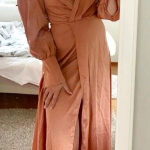 Långklänning i rostfärgad satin - Elegant långklänning i rostfärgad satin med omlott-design och långa, transparenta ärmar. Klänningen har en djup slits framtill och en vacker drapering över bysten. Aldrig använd - endast provad.
