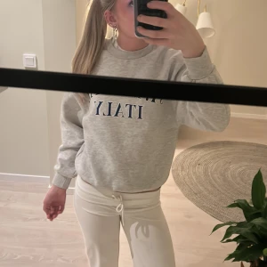 Grå sweatshirt från Gina Tricot - Säljer en grå sweatshirt från Gina Tricot med texten 'ST. MORITZ ITALY' i blått. Tröjan har långa ärmar och en rund halsringning. Perfekt för en avslappnad stil. Hör av dig vid intresse!